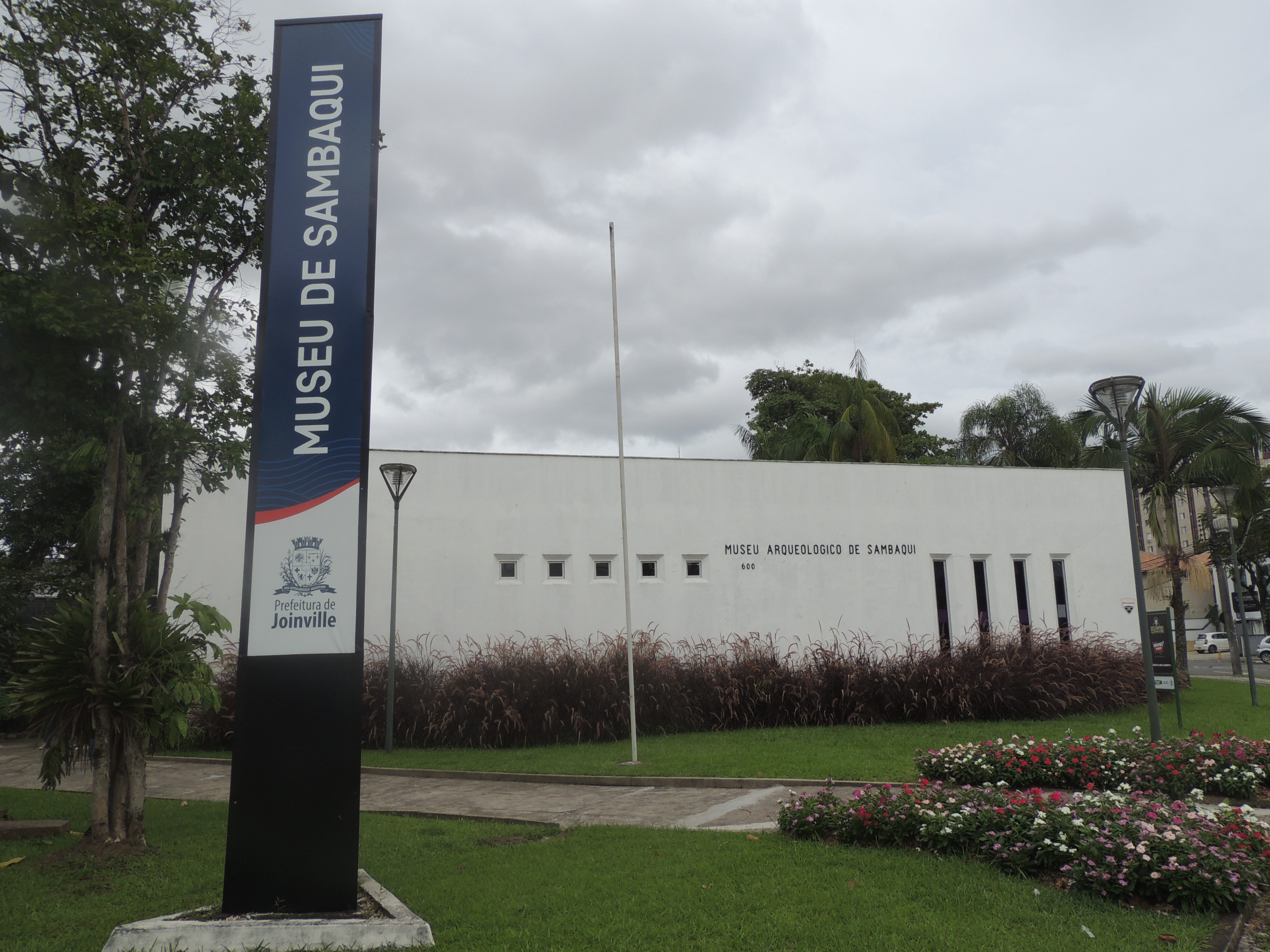 Fachada do museu arqueologico de sambaqui de Joinville. Prédio retangular branco com um totem azul com o nome do museu escrito. Em volta do prédio grama verde. O céu está branco, nublado.