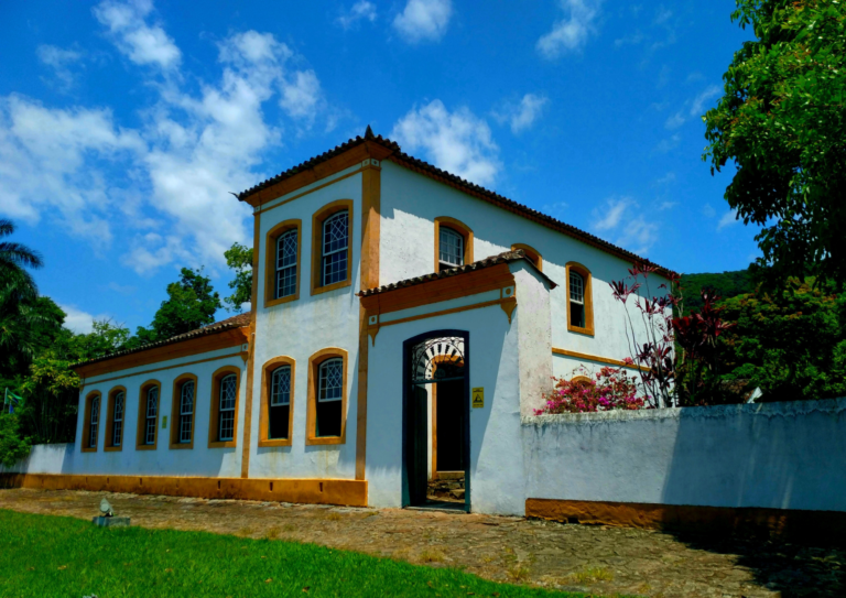 Fachada do Museu Etnográfico Casa dos Açores. Um enorme casarão colonial branco com os portais das janelas na cor amarelo ouro. O casão tem dois andares. Na fachada vê-se seis janelas embaixo e duas em cima. Também é possível ver um pequeno portão de ferro. O dia está com céu azul com nuvens brancas. Em frente ao casarão é grama verde.