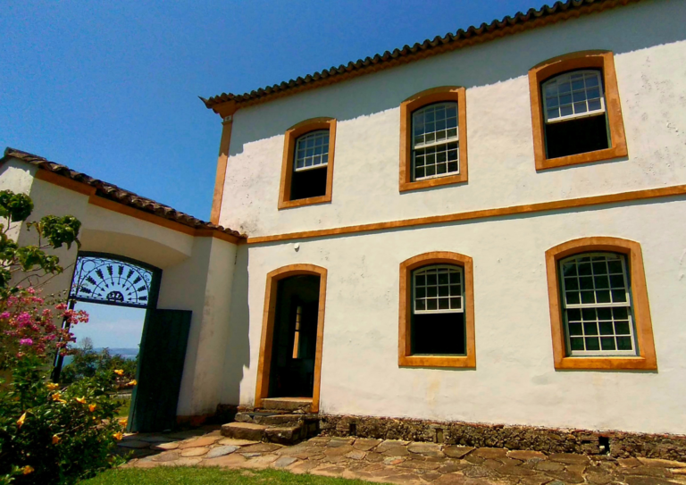 lateral sobrado com os umbrais das janelas amarelos e uma porta de madeira com portal amarelo. ao lado um portão de ferro e o fundo da imagem é o céu azul