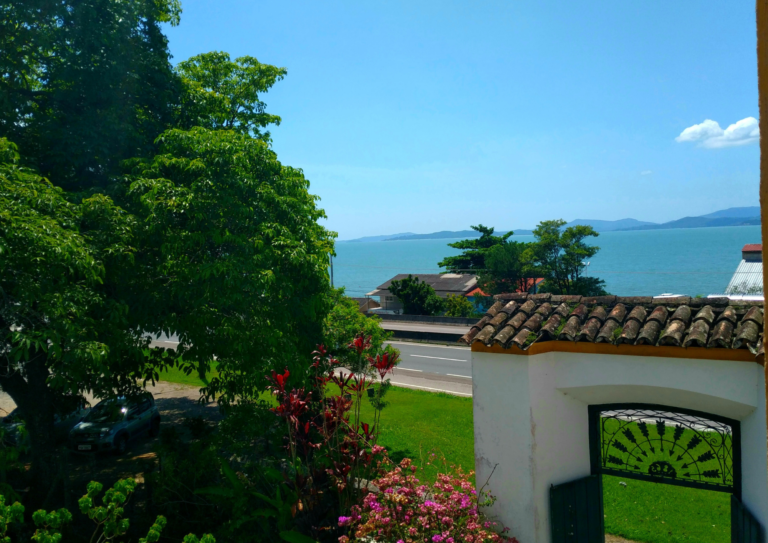 Vista do alto de dentro do sobrado. O céu azul, o mar azul contrastam com o verde da grama e da árvore