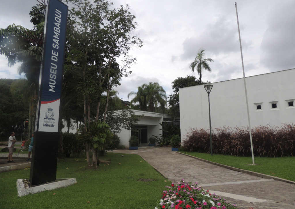 Caminho entre os dois blocos de prédios que formam a estrutura física do Museu Arqueologico de Sambaqui de Joinville. Do lado esquerdo da imagem, um totem azul com o nome do museu. Do lado direito da imagem a leteral de um prédio branco