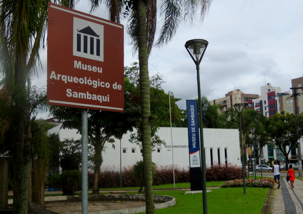 em primeiro plano uma placa marrom com o nome do museu e ao fundo a fachada do predio branco com o totem azul no meio com o nome do museu